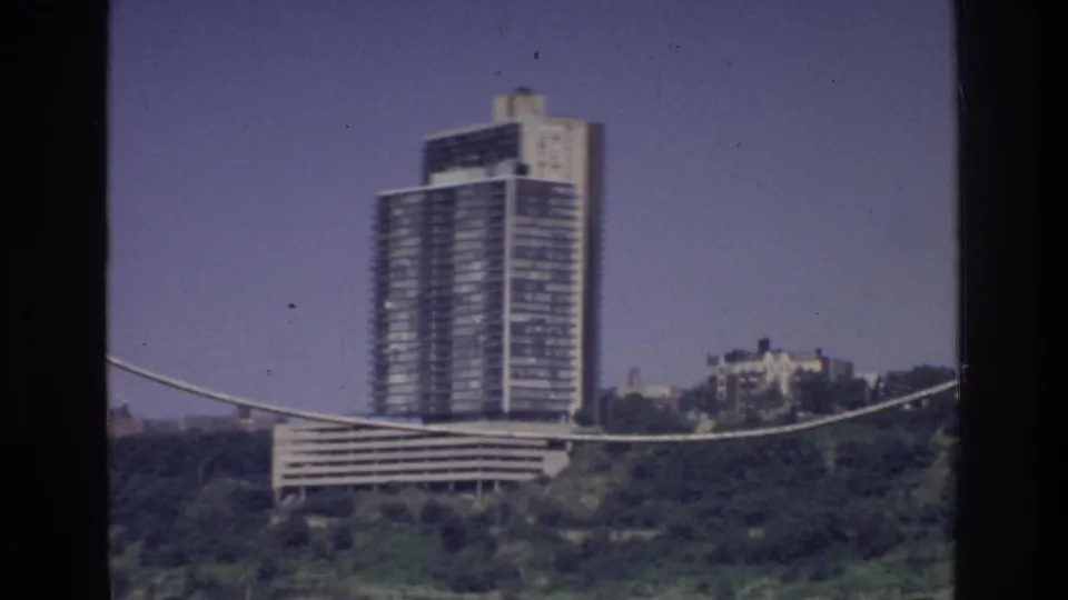 8mm film frame circa 1975, Bronx, New York — glass high-rise tower and lower stepped building seen from moving vehicle with cable in foreground.