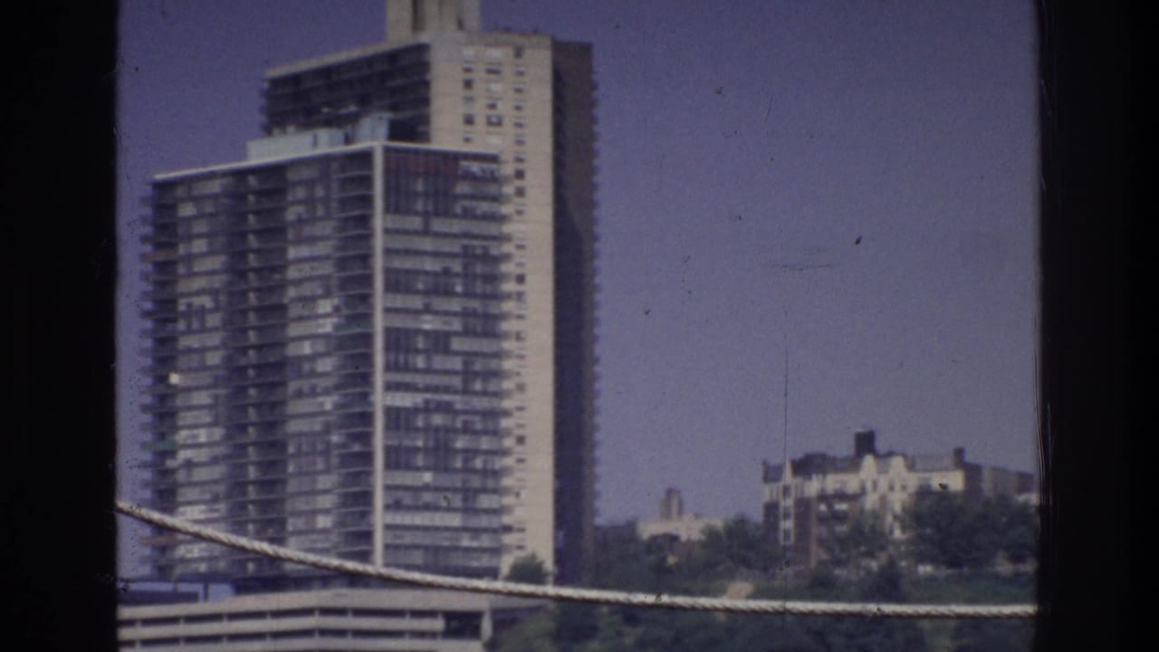 Closer view of tall glass-and-concrete tower with surrounding low-rise buildings, wooded hillside, and angled cable across lower frame.