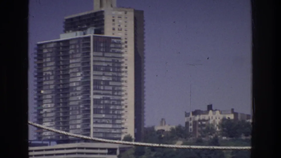 8mm frame circa 1975, Bronx, New York — modernist apartment tower fills upper frame, cable foreground, low-rise buildings and waterfront structures below.