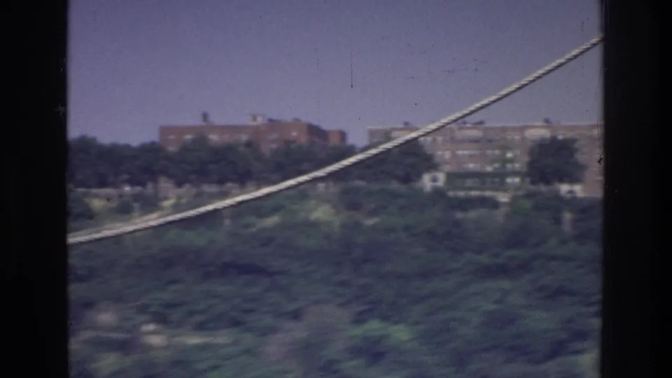 8mm frame circa 1975, Bronx or upper Manhattan waterfront — river visible in lower frame, cable diagonal, red brick residential buildings on wooded hillside.
