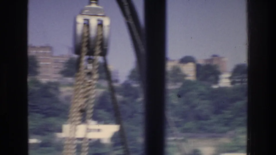 8mm frame circa 1975 — blurred cable attachment hardware fills foreground; Bronx-area red brick buildings and greenery in background.