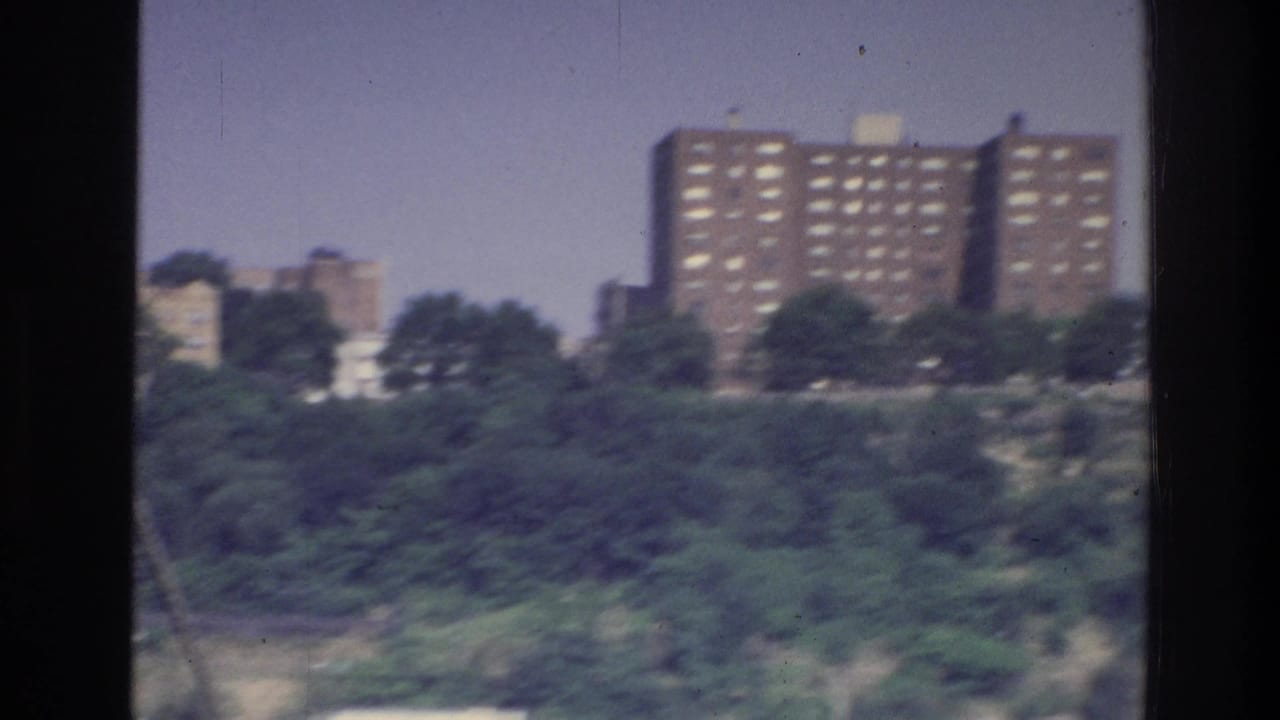 1975 boat view of a tree-covered rocky bluff topped by a large brick housing tower, consistent with a New York-area shoreline.