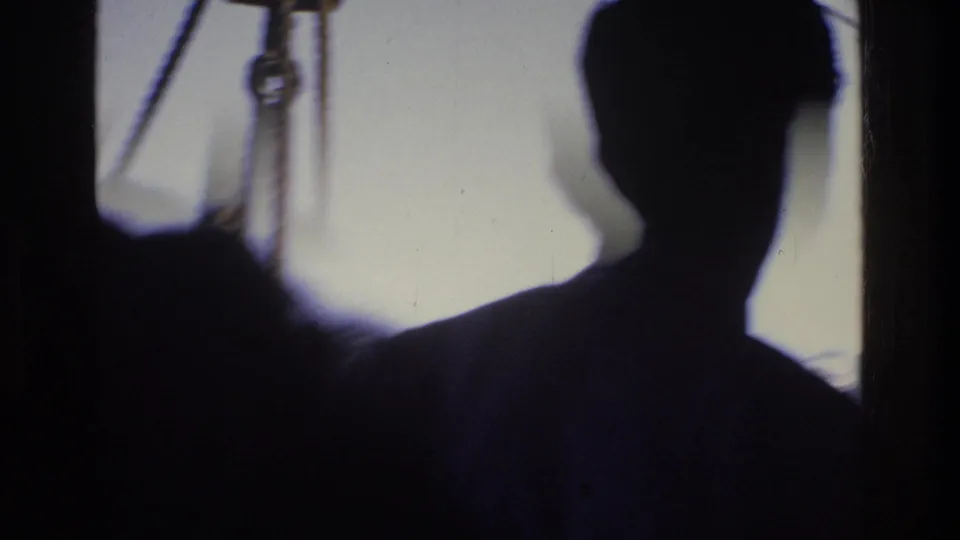 Unidentified silhouetted passenger on a 1975 boat deck, with block-and-tackle pulley hardware and rope rigging against an overexposed sky.