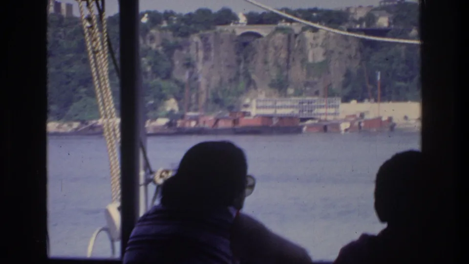 1975 view from a boat showing rocky waterfront cliffs with a stone arch and red industrial barges moored below, silhouetted passengers in the foreground.