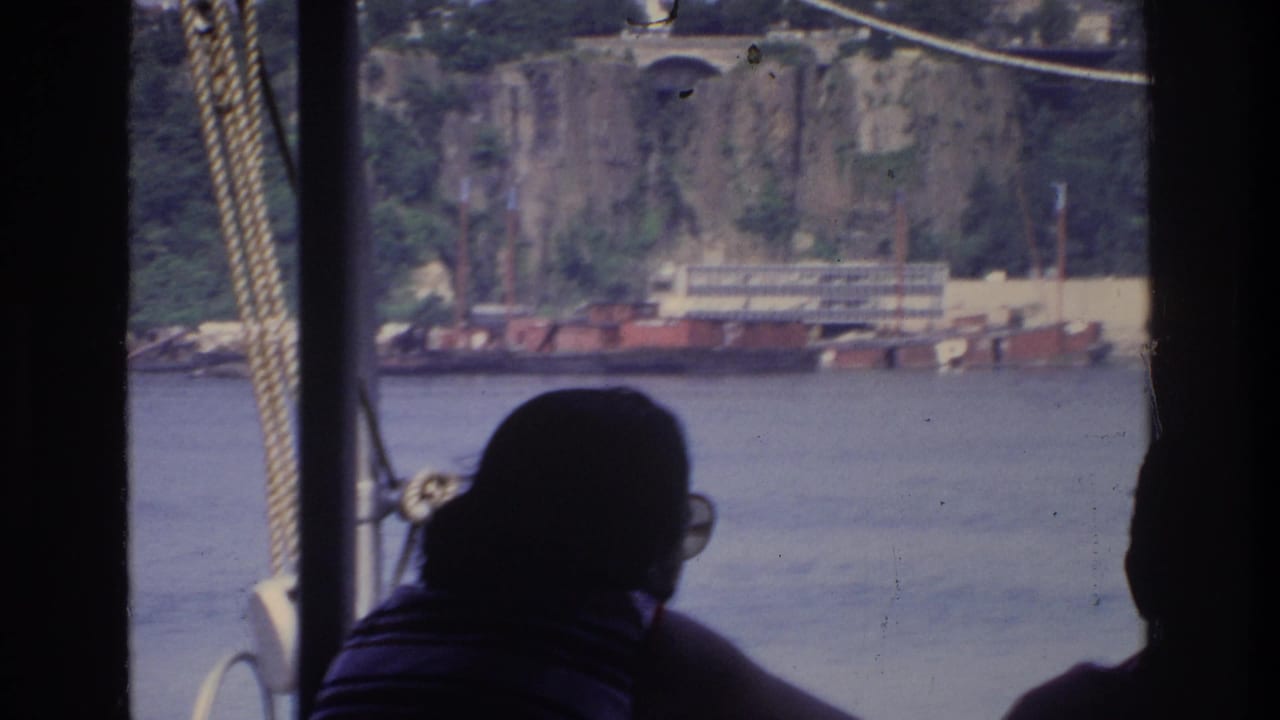 Silhouetted passenger on river boat, Bronx New York 1975; rocky cliff face, bridge arch, and industrial waterfront structures across the water.