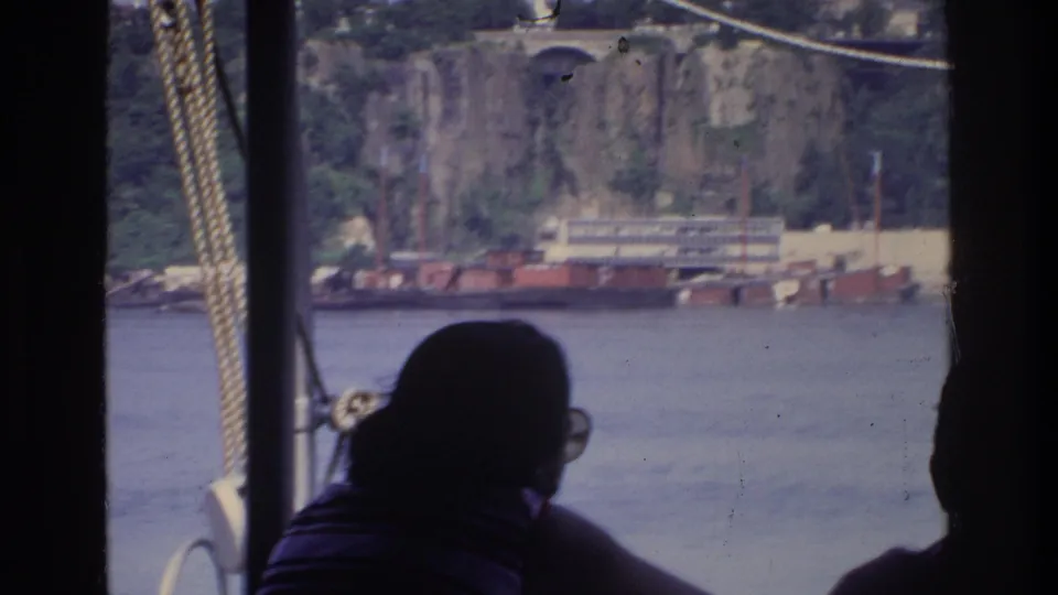 Silhouetted passenger on river boat, Bronx New York 1975; rocky cliff face, bridge arch, and industrial waterfront structures across the water.
