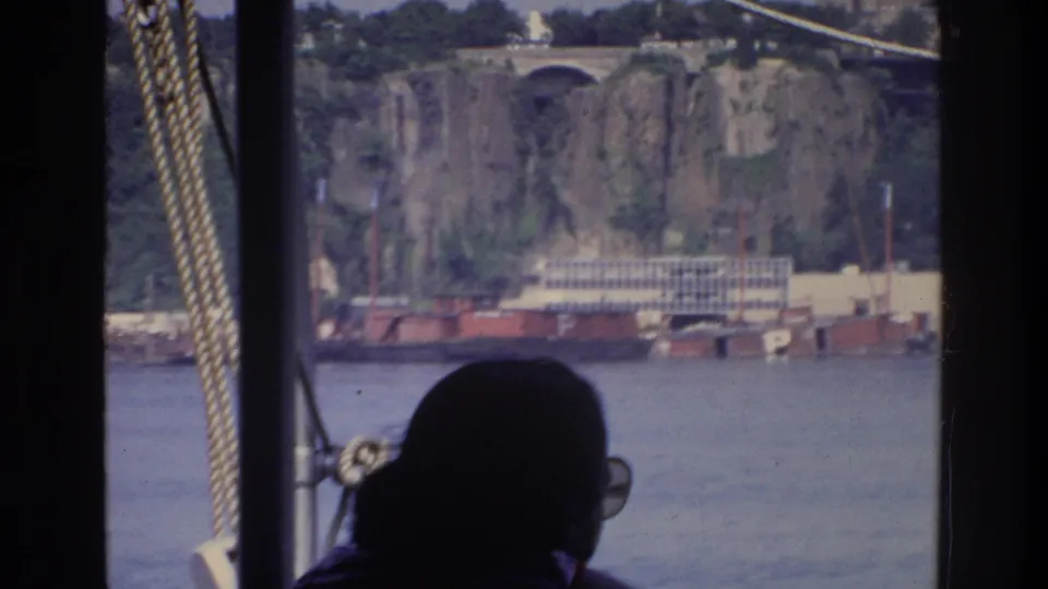 Silhouetted passenger aboard river boat, Bronx New York 1975; rocky cliff face, bridge arch, and red industrial dock structures in background.