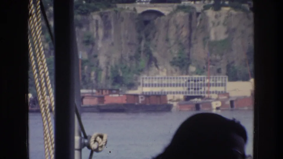 Bronx riverfront view from moving boat, 1975; rocky cliff, bridge arch, white industrial building on water, and red barges with partial letter P visible.