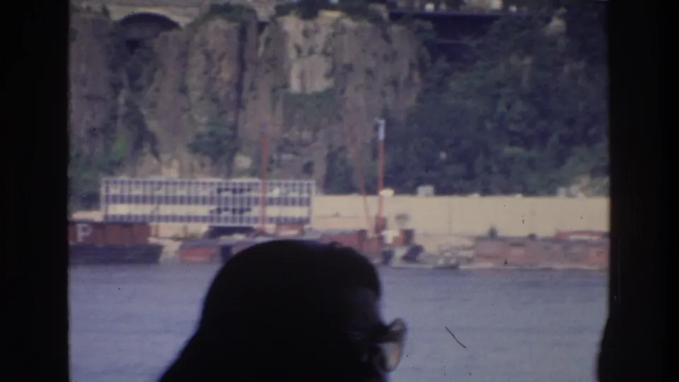Boat passenger silhouette with rocky cliff, white grid-facade building on water, and red barges along Bronx riverfront, 1975.