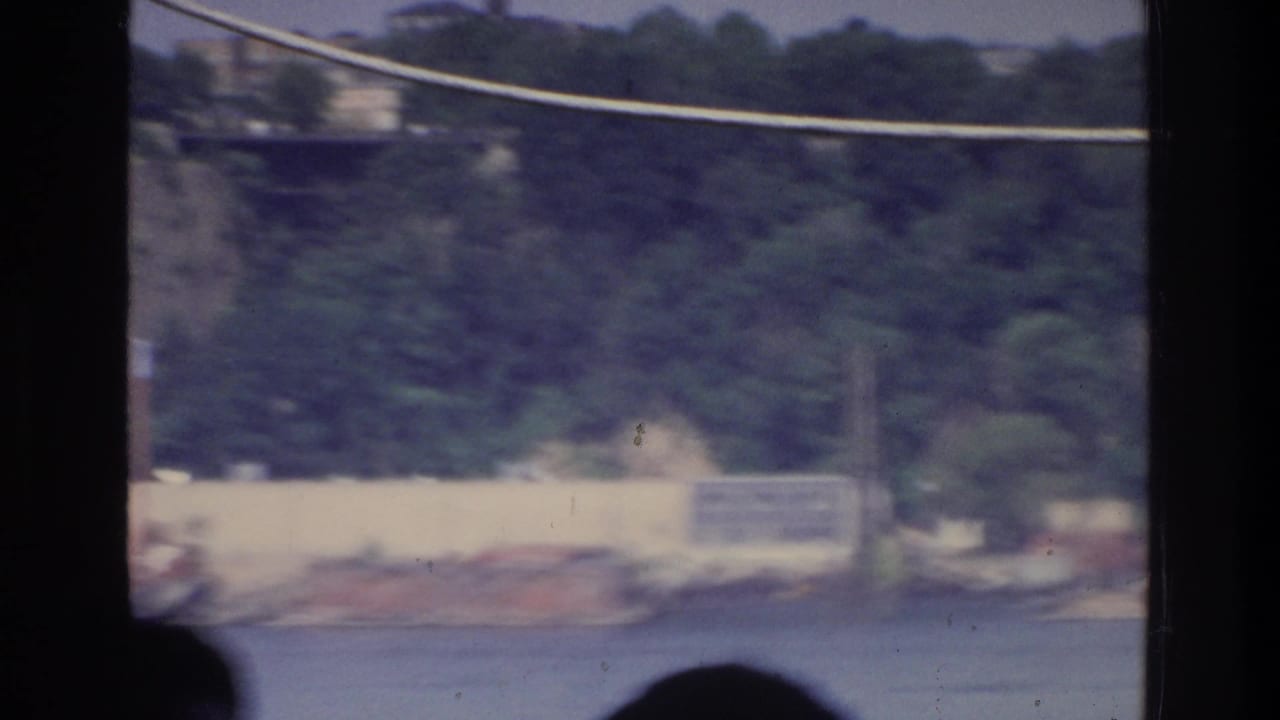 1975 8mm home movie from a boat on a New York waterway; silhouetted passengers frame a view of a wooded hillside and low waterfront structures.