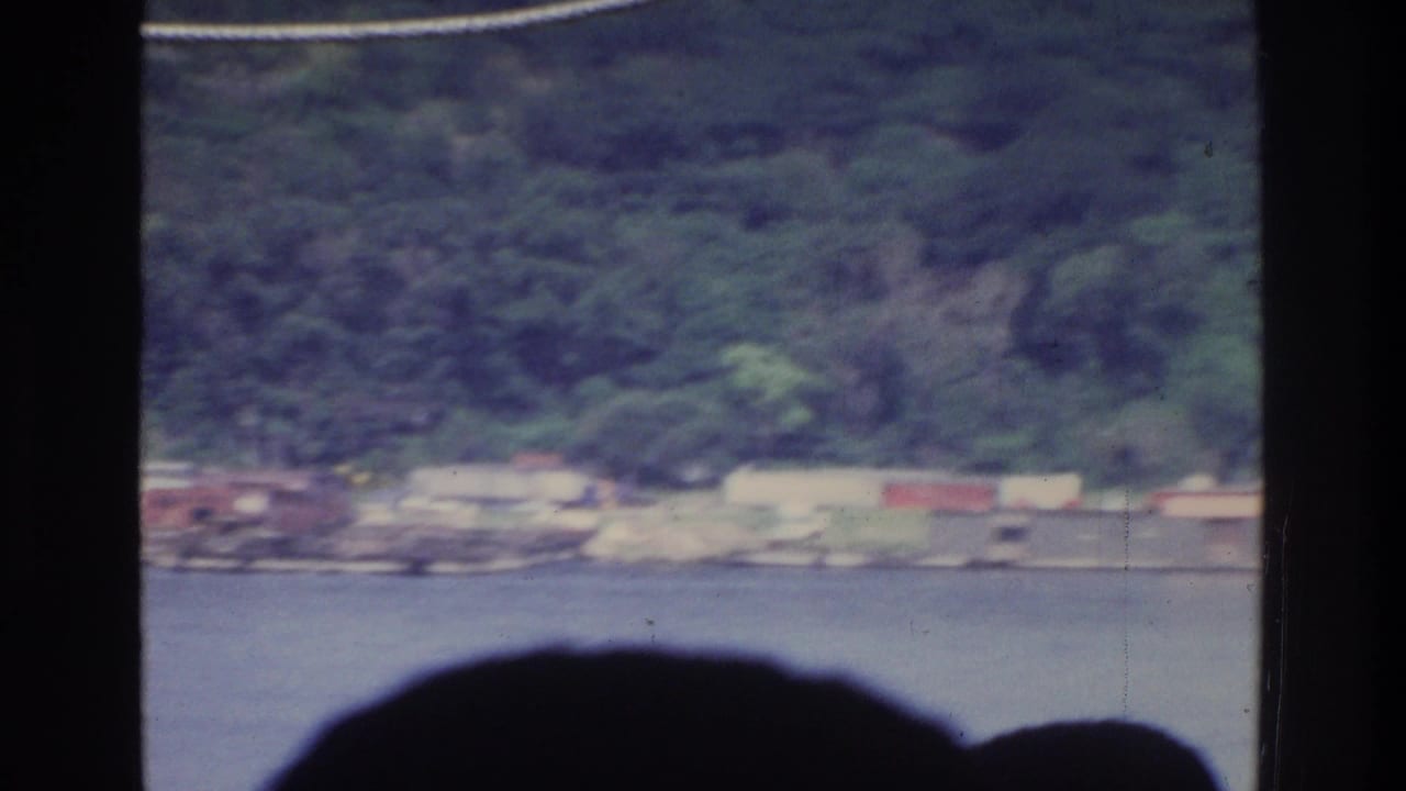 Vessel interior view looking toward a waterfront with low-rise buildings and a tree-covered ridge; silhouetted heads remain in lower foreground; white rope still visible.