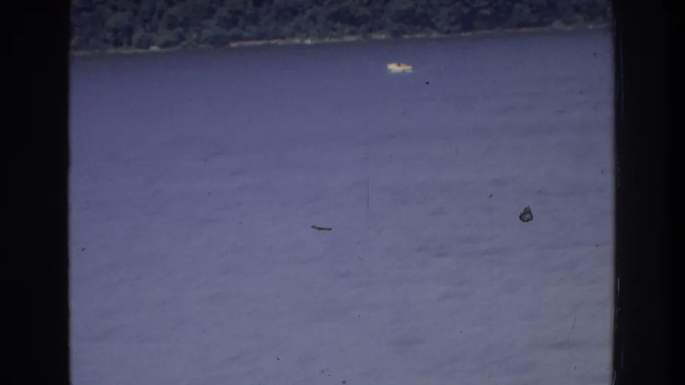 Closing 8mm frame from a New York waterway excursion, 1975; open water with a low green shoreline and small surface objects visible under aged film grain.