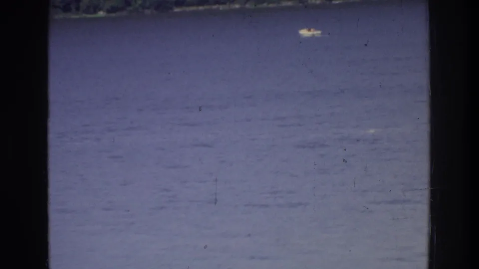 8mm home movie frame circa 1975: open water — possibly a lake or river in the New York region — with a small distant boat and tree-lined shore under a hazy sky.