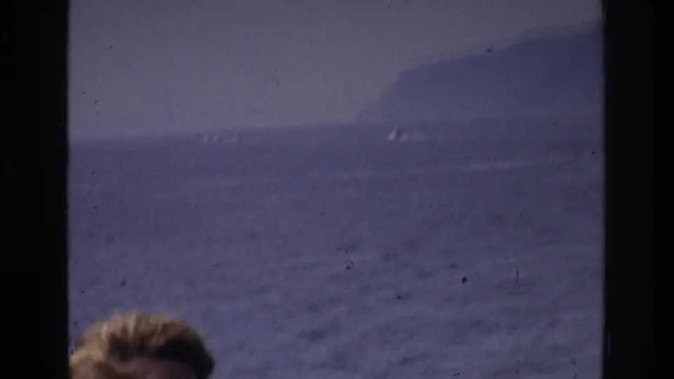 8mm home movie frame circa 1975: a person seen from behind looks out from a boat over open water, with a distant hillside or ridge visible on the horizon.