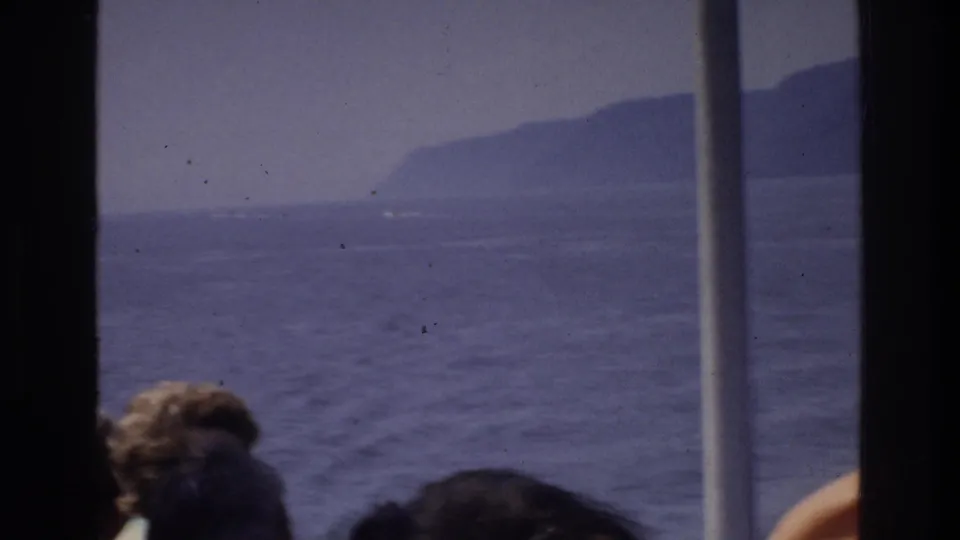 8mm home movie frame circa 1975: two boat passengers visible from behind at lower frame, a vertical mast in center, and a dark shoreline ridge across the water.