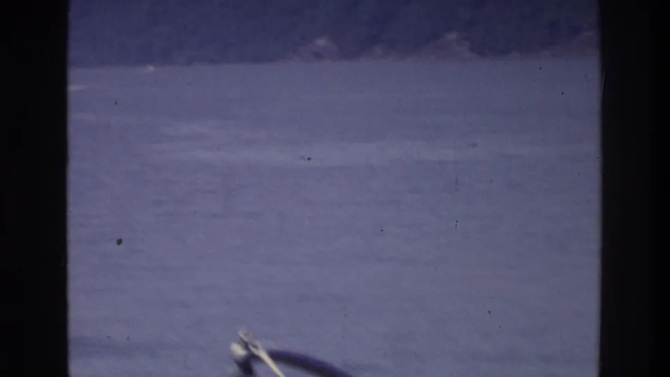 8mm home movie frame circa 1975: open water with a small boat's bow at the lower right edge and a hazy distant shoreline or hillside visible across the water.