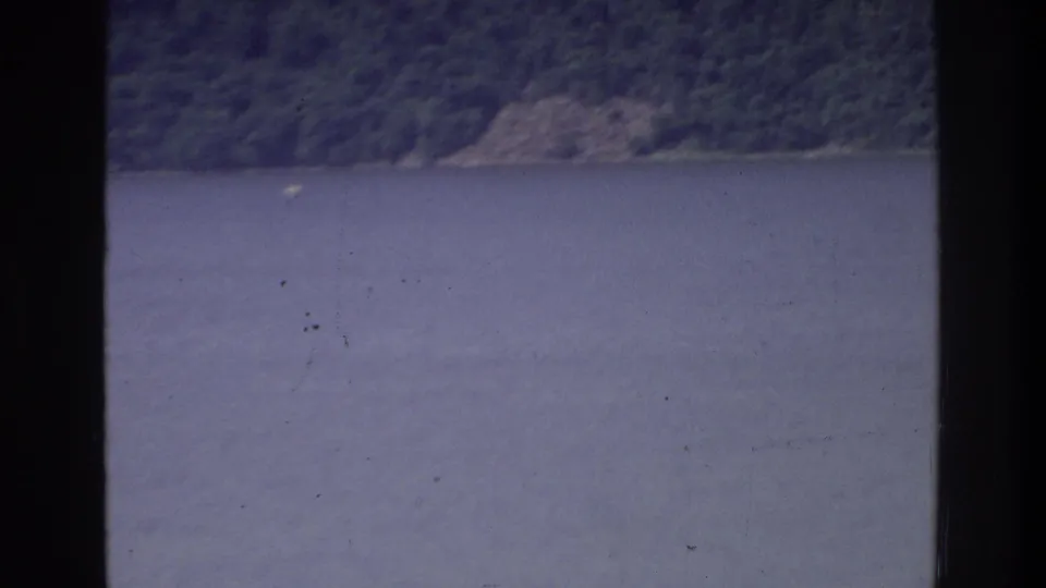 8mm home movie frame circa 1975 showing a broad water expanse and steep wooded shoreline, shot from aboard a boat on a New York-area waterway.