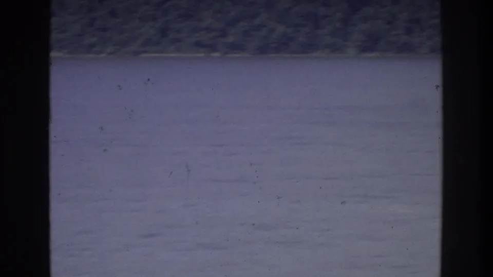 8mm home movie frame circa 1975 showing open water with a distant low shoreline, shot from a moving boat on a New York-area waterway.
