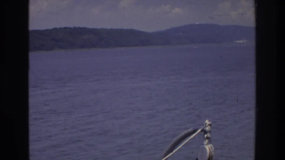 8mm home movie frame circa 1975 showing a broad waterway with distant hilly shoreline and overcast sky; a metal boat railing in the foreground confirms the on-vessel perspective.