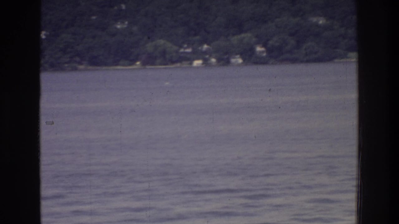 Wide river view from a tour vessel, Washington DC area, 1975; wooded shoreline with distant buildings across calm rippled water, 8mm film color fade.