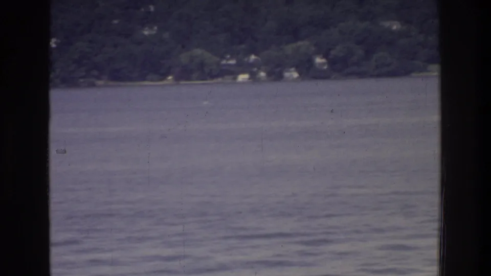 Wide river view from a tour vessel, Washington DC area, 1975; wooded shoreline with distant buildings across calm rippled water, 8mm film color fade.