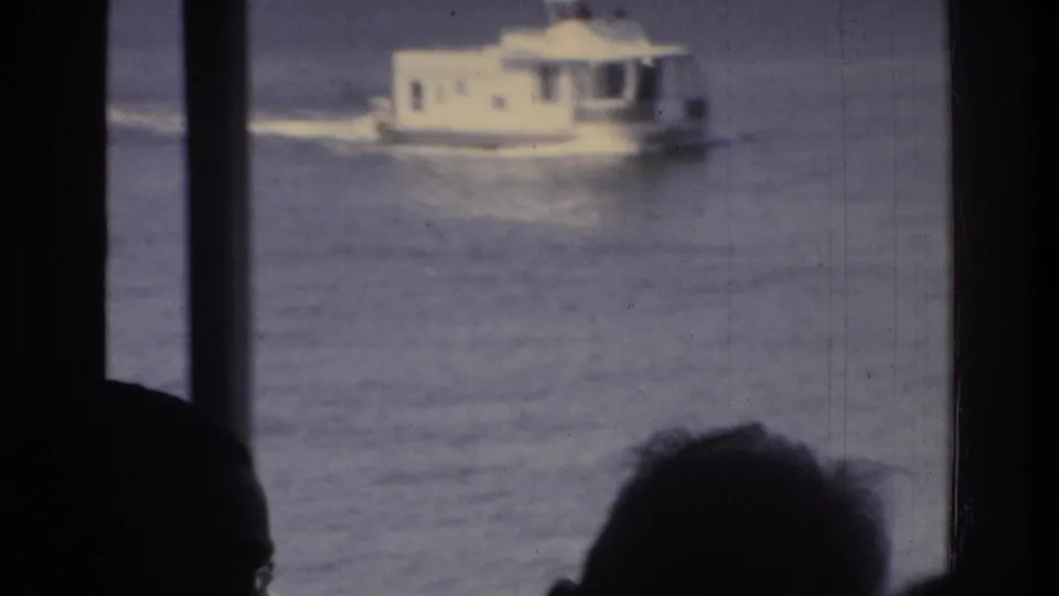 White motorboat with wake crossing river seen from a Washington DC tour boat, 1975; dark passenger silhouettes and structural columns frame the view.