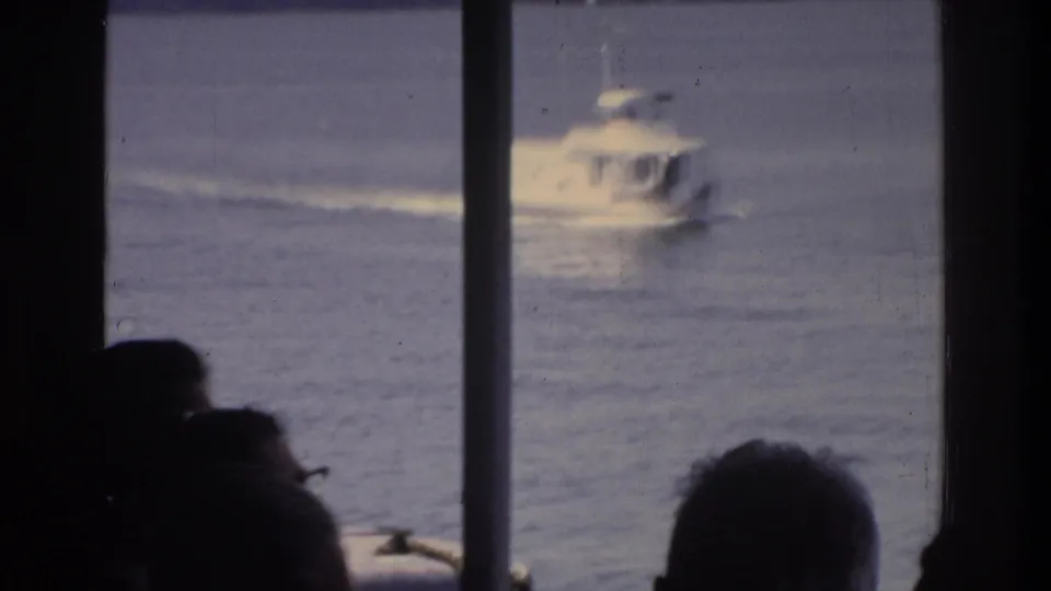 Motorboat with wake receding across river, Washington DC, 1975; viewed through tour boat structural columns over silhouetted passenger heads.