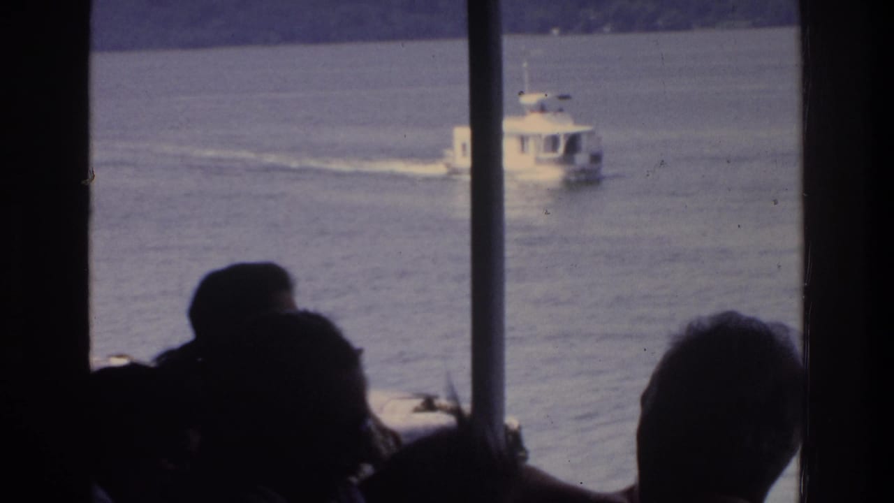 Silhouetted passengers aboard a ferry observe a white motorboat on calm lake water, 8mm home movie circa 1975.