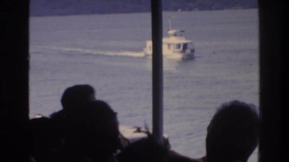 Silhouetted passengers aboard a ferry observe a white motorboat on calm lake water, 8mm home movie circa 1975.
