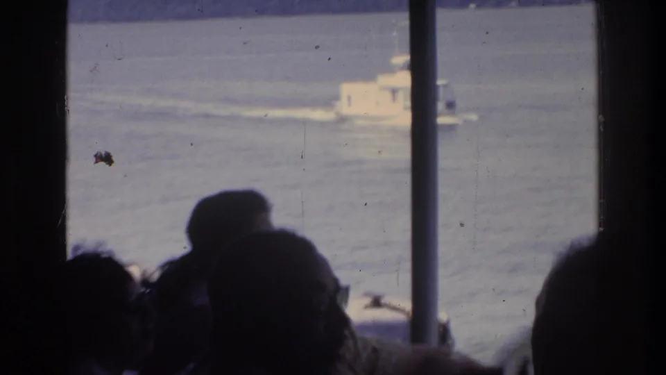 Silhouetted ferry passengers watch a white motorboat through vertical window bars; small object visible on water surface, 8mm circa 1975.