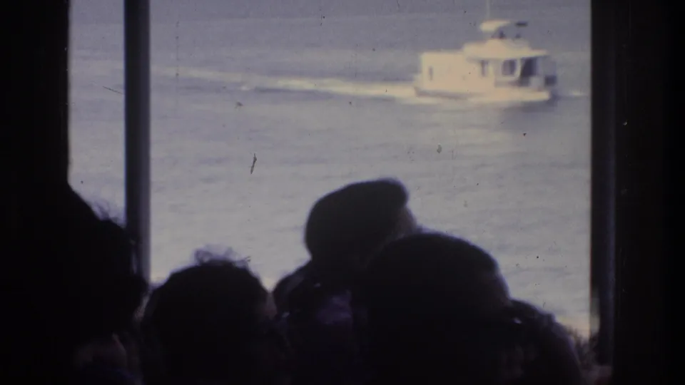 Multiple silhouetted passengers crowd a ferry window to watch a white motorboat, 8mm home movie circa 1975.