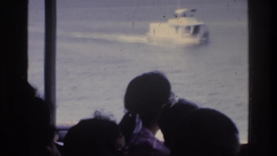 Silhouetted passengers aboard a ferry watch a white motorboat cross calm water; faint clothing color visible, 8mm home movie circa 1975.