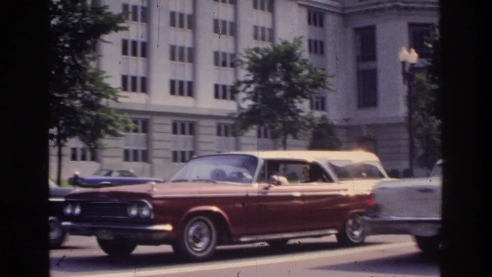 Large American cars in traffic on a Washington DC boulevard circa 1975, with classical federal-style buildings and street trees lining the road; 8mm film shot through a vehicle window.