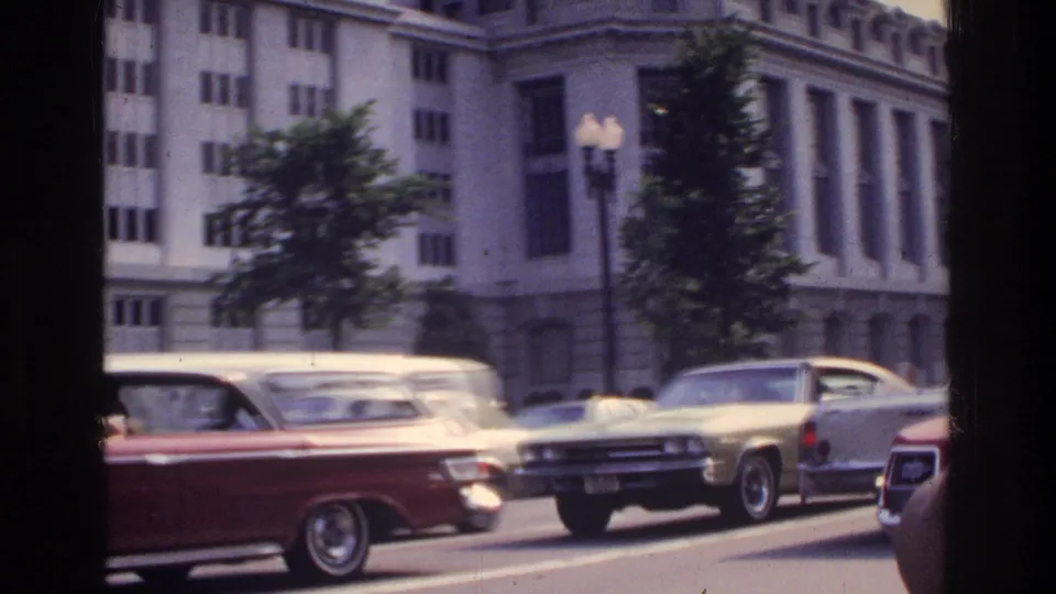 American cars in traffic on a wide Washington DC boulevard circa 1975; large columned federal-style buildings and a lamp post visible behind street trees; 8mm home movie.