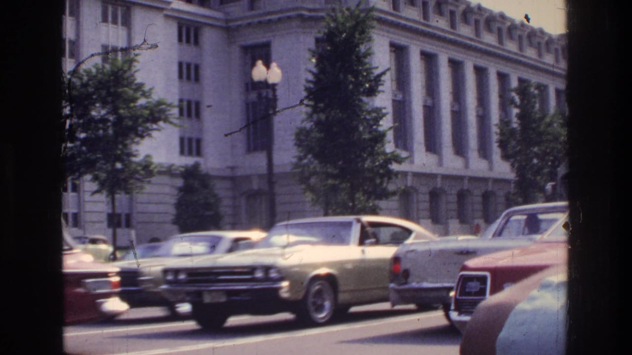 Mid-1970s street traffic including a gold coupe and red sedan passing a large neoclassical federal-style building, Washington DC, circa 1975, 8mm home movie.