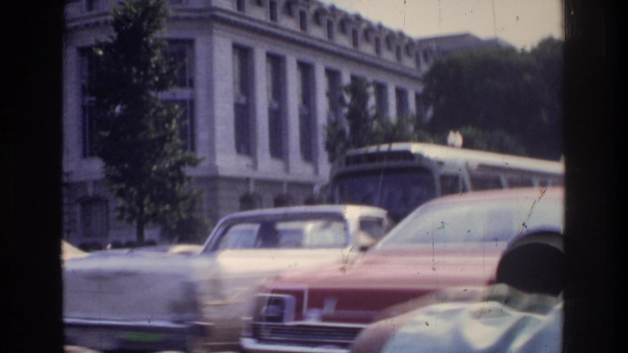 Motion-blurred large red American sedan and city transit bus moving past a neoclassical building facade, Washington DC, circa 1975, 8mm home movie.