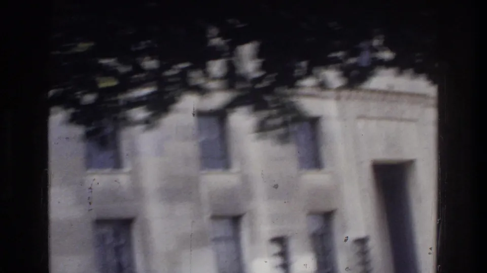 Neoclassical stone building facade with summer tree canopy overhead, motion blur, and visible film dust, Washington DC, circa 1975, 8mm home movie.