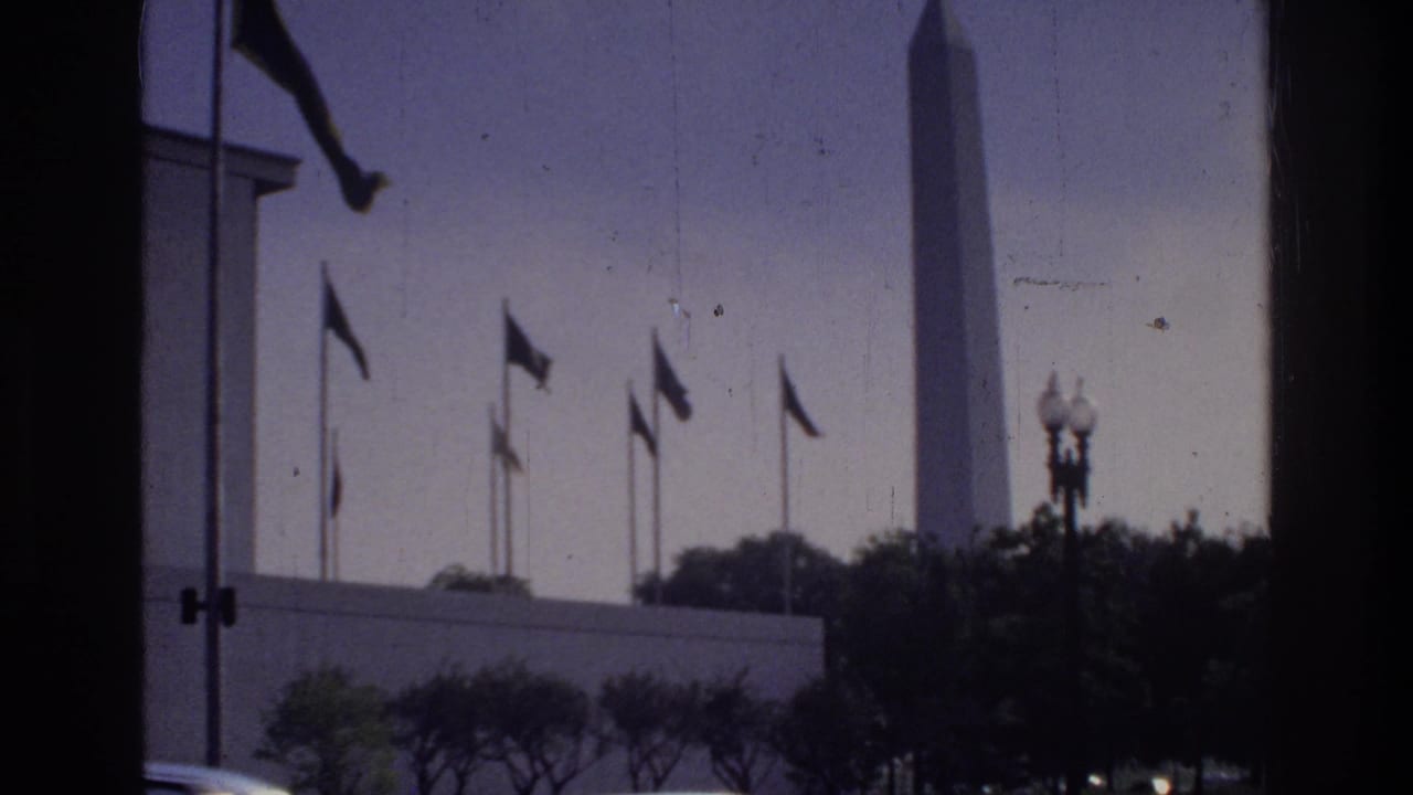 Washington Monument obelisk flanked by flag poles and globe street lamps, viewed through a window frame, Washington DC circa 1975, 8mm home movie