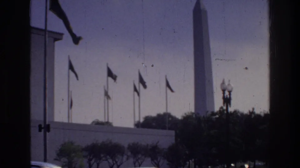 Washington Monument obelisk flanked by flag poles and globe street lamps, viewed through a window frame, Washington DC circa 1975, 8mm home movie