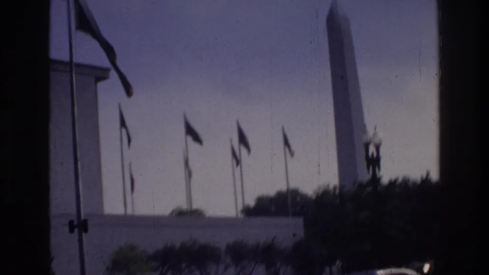 Washington Monument obelisk with row of flag poles and globe lamp cluster against pale sky, Washington DC circa 1975, 8mm home movie