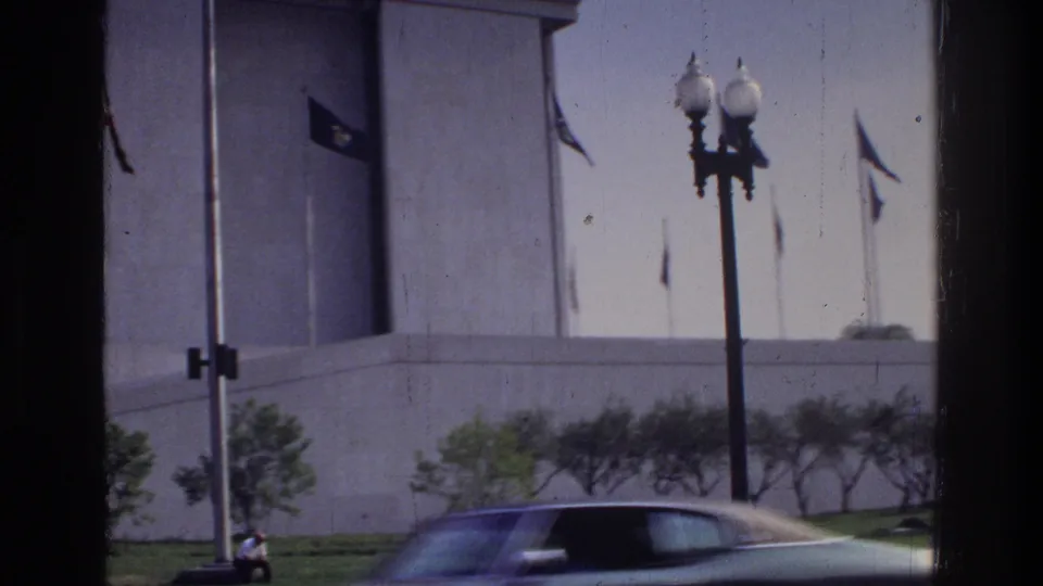 White stone wall with flags and globe street lamp, classic 1970s dark sedan in motion blur foreground, Washington DC circa 1975, 8mm
