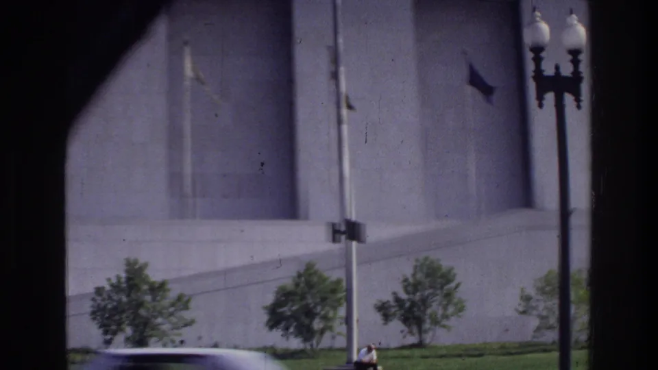 Large stone government building facade, cast-iron street lamp, and a 1970s sedan in Washington DC circa 1975, viewed through a window.