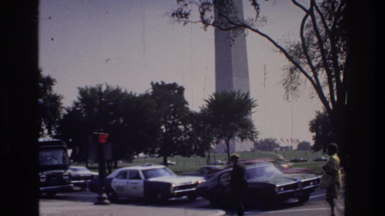 Street scene with tall obelisk in background, marked police cruiser, 1970s American sedans, bus, and pedestrians at intersection.