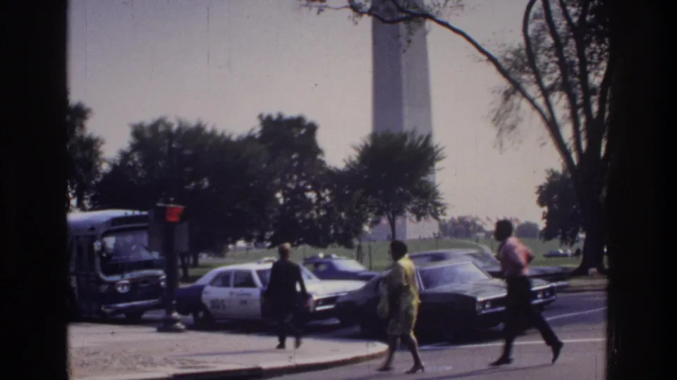Three pedestrians cross a Washington DC street circa 1975, with a marked police cruiser, 1970s sedans, a bus, and the Washington Monument rising behind them.
