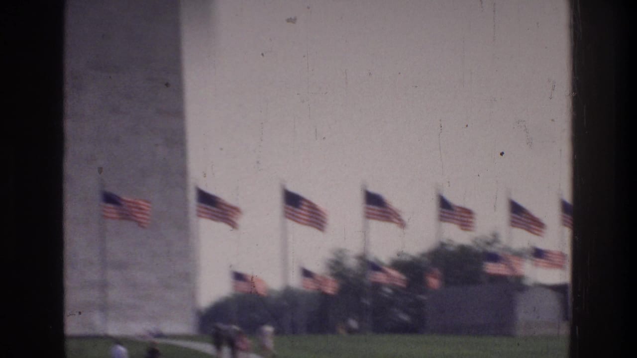 8mm home movie frame circa 1975 showing the lower section of the Washington Monument with a ring of American flags and green lawn at the base