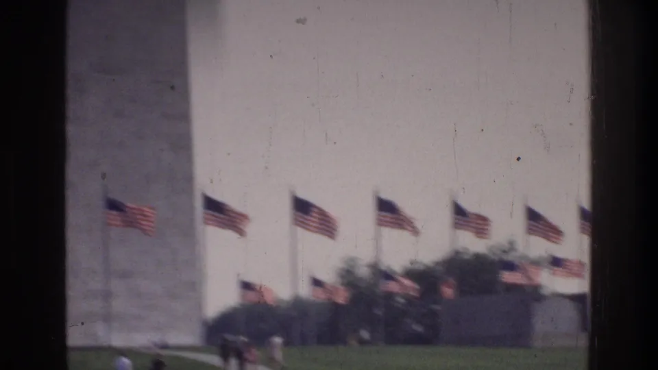 8mm home movie frame circa 1975 showing the lower section of the Washington Monument with a ring of American flags and green lawn at the base