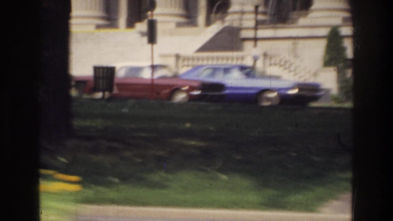 Red and blue large-body American sedans on a Washington DC street, circa 1975, with a neoclassical government building visible in the background.