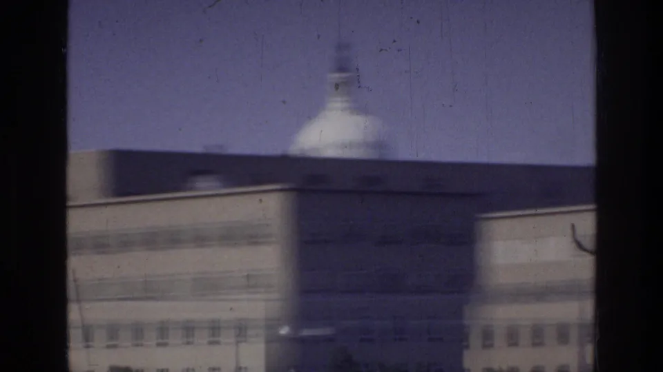 A white dome — possibly the US Capitol — rises above a rectangular federal building in Washington DC, circa 1975, filmed from a moving vehicle.