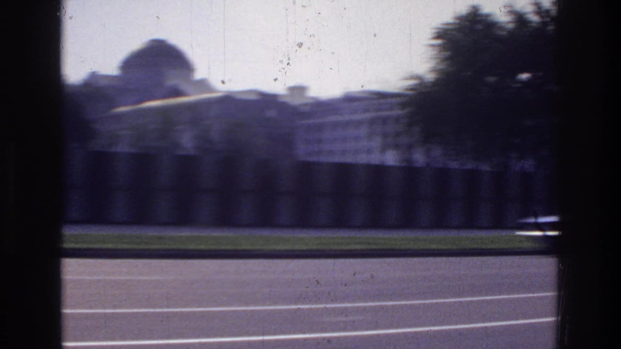 Motion-blurred POV car window shot showing a domed structure and a long neoclassical building facade with a green lawn strip below.