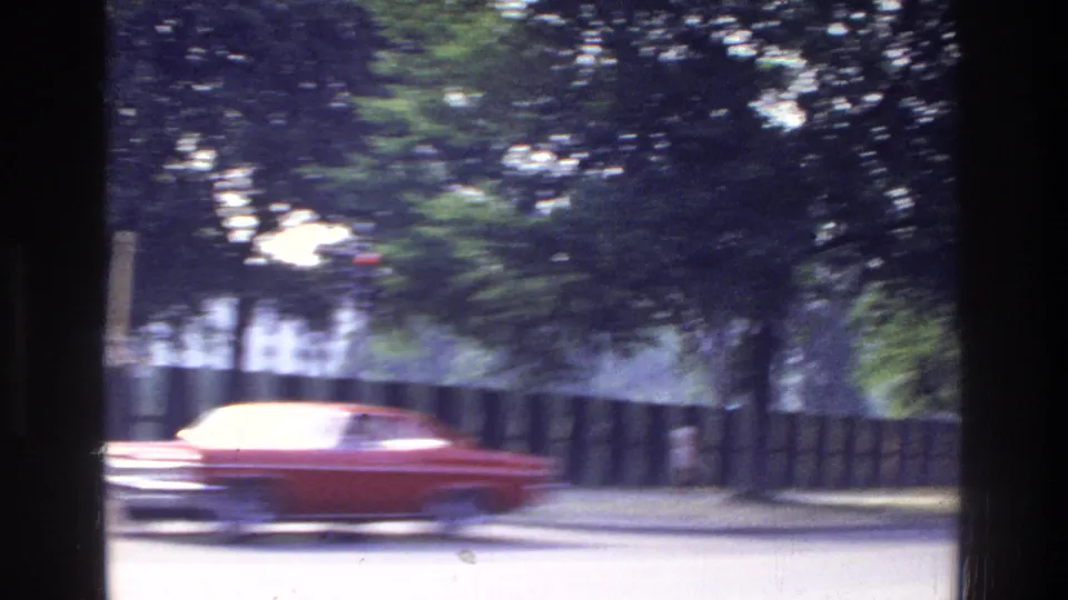 8mm home movie of a red 1960s–70s American car driving along a tree-lined Washington DC boulevard in 1975, with dappled sunlight filtering through mature foliage.